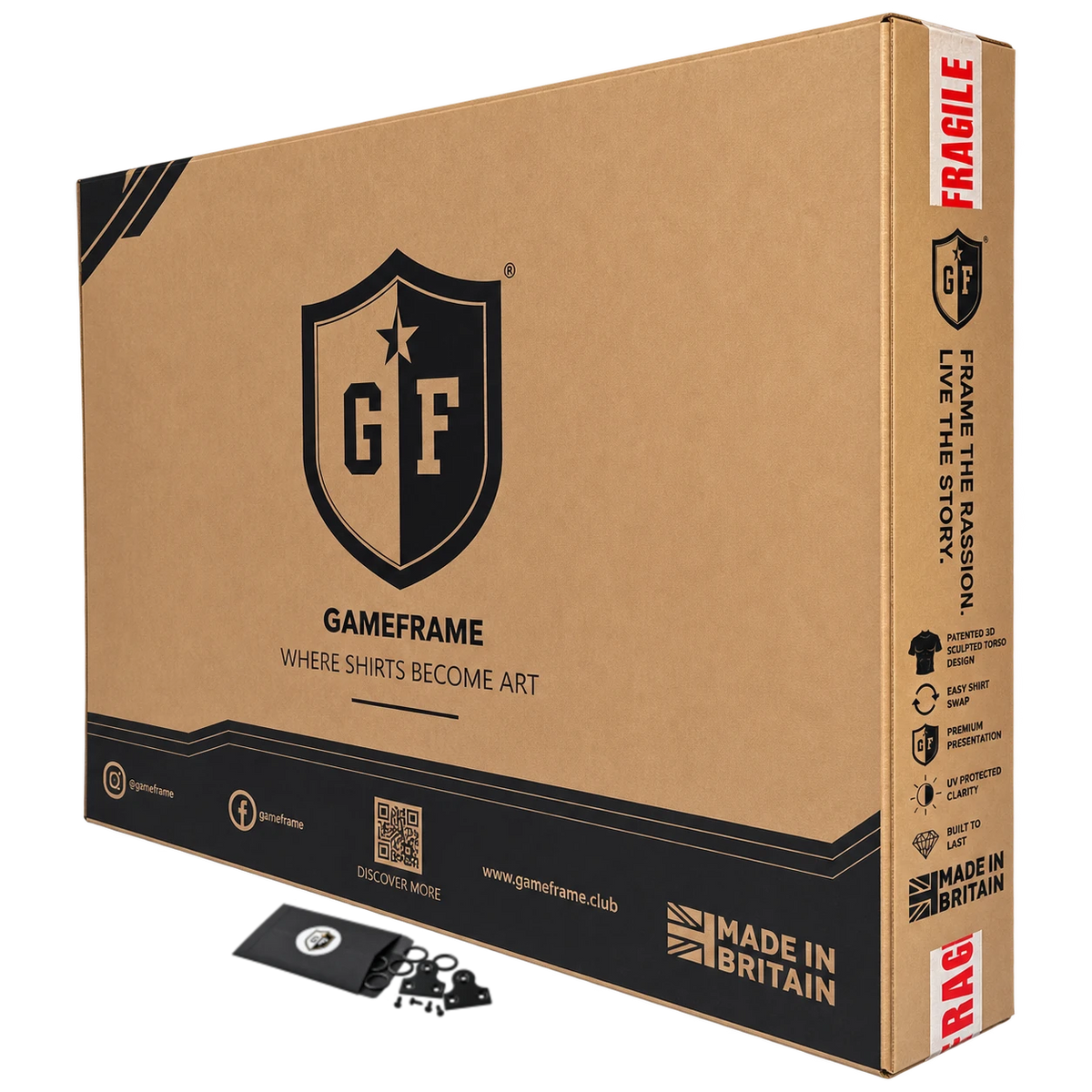 A large brown cardboard product box for Gameframe, shown at an angle with a small black accessory piece and a round black-and-white tag placed in front. The box features the Gameframe shield logo, 'GAMEFRAME' text, and 'MADE IN BRITAIN' branding on a clean white background.